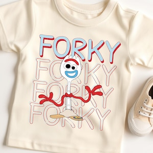 May include: Cream-coloured t-shirt with the word "FORKY" repeated, and a cartoon fork character. The fork has a smiling face, red arms, and stands on a wooden base. A pair of beige trainers are visible on the right.