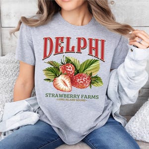 a woman sitting on a bed wearing a delphi strawberry farms shirt