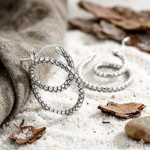 May include: A pair of silver hoop earrings featuring a textured snake scale pattern. One earring is positioned upright, showing a snake head detail at the clasp, while the other earring lies flat, displaying a continuous circular band of scales.