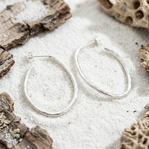 May include: A pair of silver hoop earrings with a textured, hammered finish. The earrings feature an elongated, oval shape and include standard post-back closures. The metal surface has a reflective, polished appearance with subtle indentations.