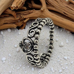 May include: Silver ring with a heart-shaped design and a small, clear gemstone. The ring features intricate scrollwork and a beaded band. The ring is displayed on a sandy surface with driftwood in the background.