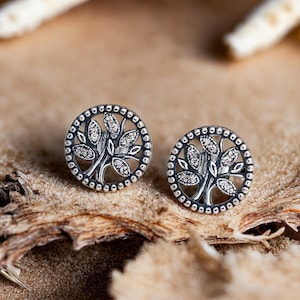 May include: A pair of silver earrings featuring a tree of life design. The earrings are round and have a delicate, intricate design.