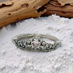 May include: A silver ring featuring a large, clear central gemstone, flanked by smaller stones. The ring rests on white sand, with driftwood in the background. The band of the ring has a decorative pattern. The ring's band has a decorative design.