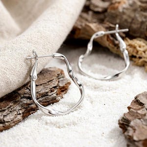 May include: A pair of silver-toned hoop earrings featuring an irregular, wavy, and organic shape. The earrings have a polished metallic finish and a hinged post closure mechanism, designed with a continuous, flowing silhouette that lacks a perfect circle.