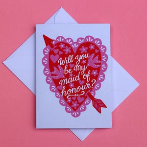 May include: A white greeting card with a red heart illustration featuring a pink scalloped border, small flowers, and two birds. A red arrow pierces the heart, which displays the white text: Will you be my maid of honour? A white envelope is behind the card.