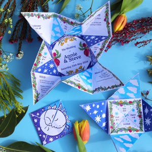 May include: A blue and white wedding invitation in the shape of a star. The invitation is folded and has a floral design. The text on the invitation reads "The Wedding of Annie & Steve, Saturday 25th May 2014".