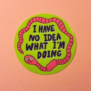 May include: A circular sticker featuring a green background with a pink worm illustration. The sticker has the text "I HAVE NO IDEA WHAT I'M DOING" in blue. The sticker is set against a pink backdrop.