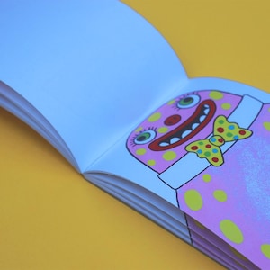 Peut inclure: Carnet de croquis ouvert avec une illustration de clown de dessin animé. Le clown a un visage rose avec des pois jaunes, un nez rouge et un nœud papillon jaune et vert. Le fond est jaune.