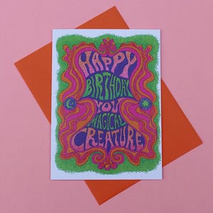 May include: A birthday card with the words "HAPPY BIRTHDAY YOU MAGICAL CREATURE" in a colourful, psychedelic design. The card is on an orange envelope, set against a pink background.