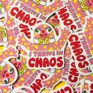 Könnte beinhalten: Eine Ansammlung von rosa Aufklebern mit einem Cartoon-Charakter mit einem lächelnden Gesicht, grünen Augen und einer gelben Fliege. Der Text "I THRIVE IN CHAOS" ist rot.
