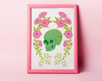 Può includere: Una cornice rettangolare rosa contenente una stampa bianca con un teschio verde centrale circondato da due steli simmetrici di fiori rosa con foglie verdi e radici sottili e ramificate nella parte inferiore della composizione.