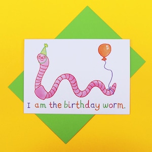 May include: A birthday card with a cartoon pink worm wearing a party hat and holding an orange balloon. The words "I am the birthday worm" are written below. The card is on a green envelope, set against a yellow background.