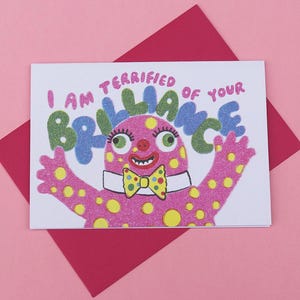 May include: A greeting card featuring a pink monster with yellow spots, a bow tie, and the text "I AM TERRIFIED OF YOUR BRILLIANCE". The card is on a red envelope, set against a pink background. The monster has green eyes.