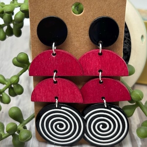 May include: Dangle earrings featuring a black circular stud, two stacked red wooden semi-circles, and a bottom black circular disc with a white spiral pattern. The components are connected by silver-toned metal jump rings on a brown display card.