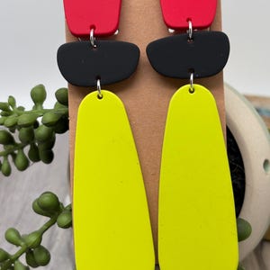 May include: A pair of dangle earrings featuring three stacked geometric shapes in matte red, black, and neon yellow. The top piece is a red square with rounded corners, the middle is a black horizontal oval, and the bottom is a long yellow teardrop.