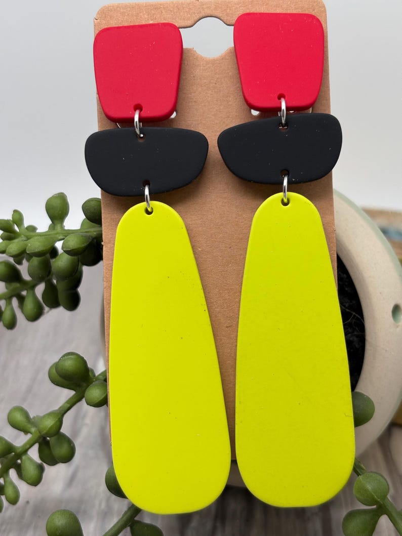 May include: A pair of dangle earrings featuring three stacked geometric shapes in matte red, black, and neon yellow. The top piece is a red square with rounded corners, the middle is a black horizontal oval, and the bottom is a long yellow teardrop.