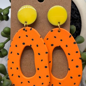 May include: A pair of dangle earrings featuring a bright yellow circular stud top connected to a large, irregular, orange oval-shaped pendant with a central cutout. The orange pendants are decorated with small, scattered black polka dots.