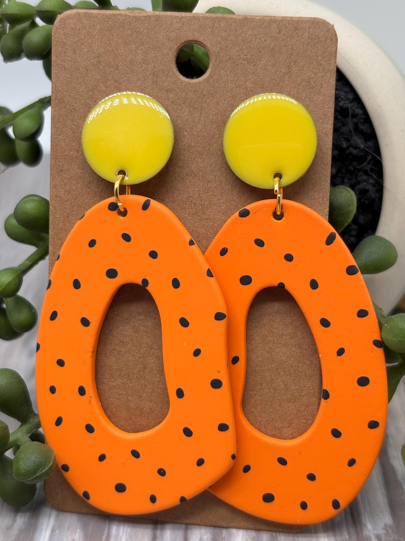 May include: A pair of dangle earrings featuring a bright yellow circular stud top connected to a large, irregular, orange oval-shaped pendant with a central cutout. The orange pendants are decorated with small, scattered black polka dots.