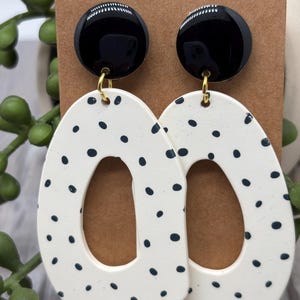 May include: A pair of dangle earrings featuring a glossy black circular stud connected to a large, off-white, irregular oval-shaped pendant with a central cutout. The off-white pendants are decorated with a pattern of small, scattered black dots.