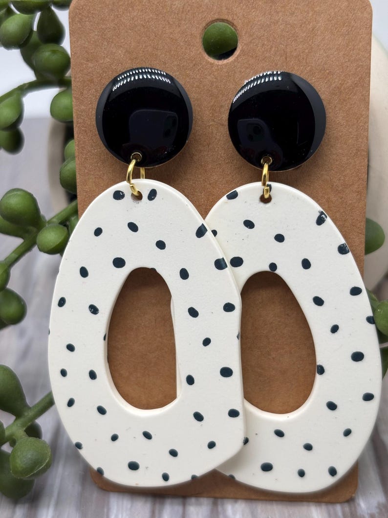 May include: A pair of dangle earrings featuring a glossy black circular stud connected to a large, off-white, irregular oval-shaped pendant with a central cutout. The off-white pendants are decorated with a pattern of small, scattered black dots.