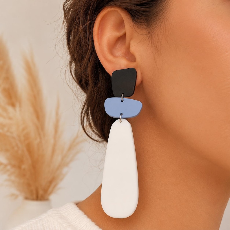 May include: A drop earring featuring three stacked geometric shapes in black, light blue, and white. The top piece is a small black trapezoid, the middle is a light blue rounded rectangle, and the bottom is a large, elongated white teardrop shape.