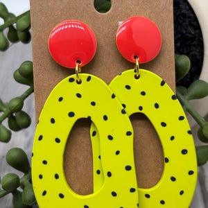 May include: A pair of dangle earrings featuring a bright red circular stud at the top connected to a large, irregular oval-shaped drop in neon yellow with a black polka dot pattern. The earrings are displayed on a brown rectangular card.