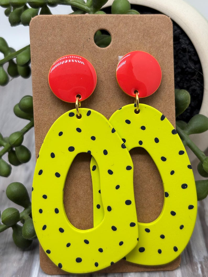 May include: A pair of dangle earrings featuring a bright red circular stud at the top connected to a large, irregular oval-shaped drop in neon yellow with a black polka dot pattern. The earrings are displayed on a brown rectangular card.