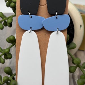 May include: A pair of dangle earrings featuring three stacked geometric shapes in black, light blue, and white. The top piece is a black square, the middle is a light blue rounded rectangle, and the bottom is a long white teardrop shape.