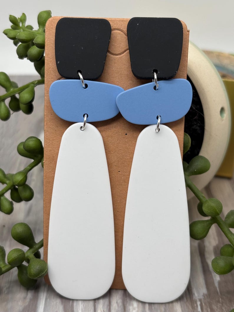 May include: A pair of dangle earrings featuring three stacked geometric shapes in black, light blue, and white. The top piece is a black square, the middle is a light blue rounded rectangle, and the bottom is a long white teardrop shape.