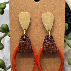 May include: Earrings with a teardrop-shaped gold-tone top, brown leather accent, and a bright orange oval hoop. The earrings are displayed on a brown card.