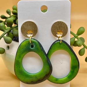 Pode incluir: Um par de brincos de resina verde com um detalhe em metal dourado. Os brincos têm a forma oval com um círculo menor cortado no centro.