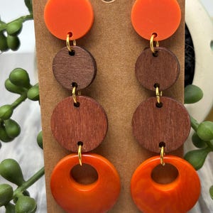 Pode incluir: Um par de brincos pendentes com quatro componentes circulares empilhados. O disco superior é laranja sólido, seguido por dois discos de madeira castanho-escuro e um anel laranja na parte inferior. As peças estão ligadas por pequenos anéis metálicos dourados.