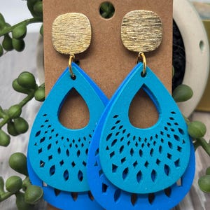 May include: A pair of blue teardrop earrings with a gold square stud. The earrings have a layered design with cut-out patterns. The top layer is a lighter shade of blue, and the bottom layer is a darker shade of blue.