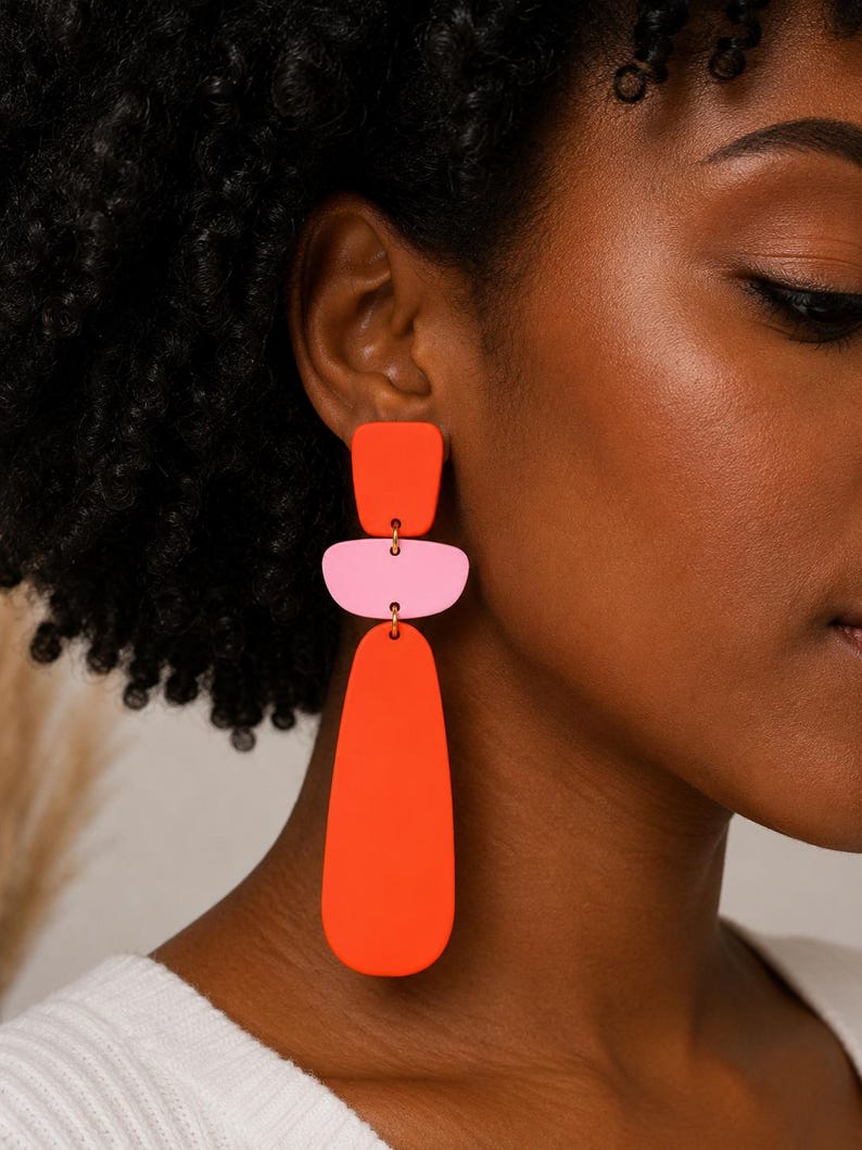 May include: A pair of drop earrings featuring three stacked geometric shapes in matte finish. The top piece is a small orange square, the middle is a light pink rounded rectangle, and the bottom is a large, elongated orange teardrop shape.