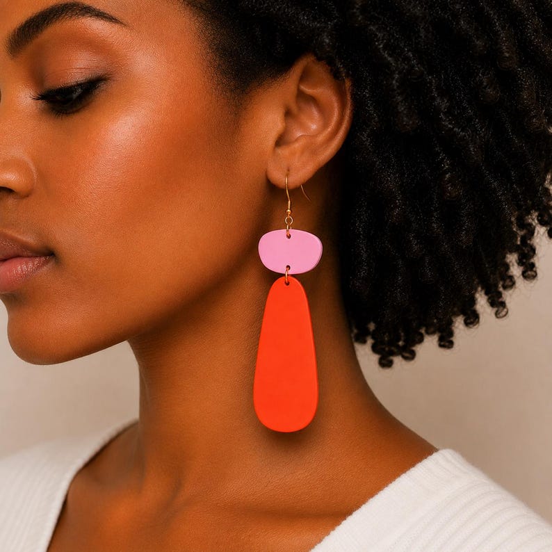May include: A pair of drop earrings featuring a small, rounded pink top piece connected to a larger, elongated, teardrop-shaped orange bottom piece. The earrings hang from thin gold-toned metal hooks, creating a two-tiered, colorful design.
