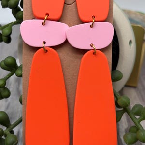 May include: A pair of dangle earrings featuring three stacked geometric shapes in bright orange and light pink. The top piece is a small square, the middle is a rounded horizontal rectangle, and the bottom is a long, smooth teardrop shape.