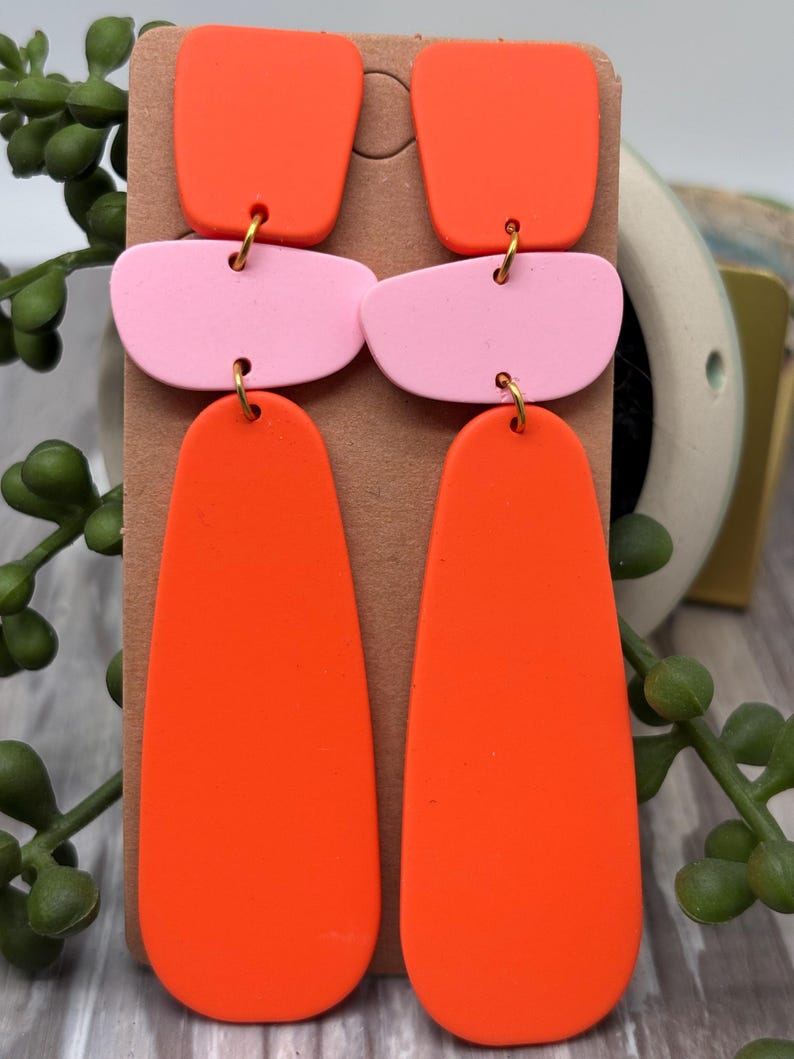 May include: A pair of dangle earrings featuring three stacked geometric shapes in bright orange and light pink. The top piece is a small square, the middle is a rounded horizontal rectangle, and the bottom is a long, smooth teardrop shape.