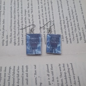 May include: A pair of silver earrings with a book charm. The book covers are blue and white with the title "Six of Crows" printed on the front.