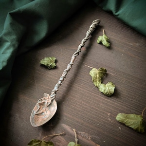 Puede incluir: Una cuchara de metal hecha a mano con un mango retorcido y un cuenco en forma de hoja. La cuchara tiene un aspecto rústico y antiguo, con un diseño detallado. La cuchara se exhibe sobre una superficie de madera oscura con hojas de hiedra secas esparcidas.