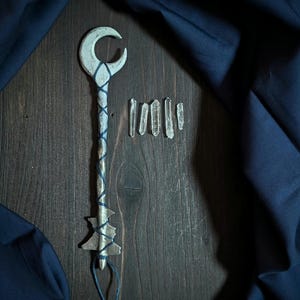 Puede incluir: Una varita mágica plateada en forma de llave con una luna creciente en la parte superior y un mango decorativo envuelto en hilo azul. Varios cristales de cuarzo transparente están dispuestos junto a la varita sobre una superficie de madera oscura. Un paño azul oscuro está drapeado en el fondo.