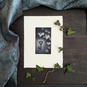 May include: A black and white print of a key surrounded by leaves. The artwork is on a white paper with a deckled edge. Green ivy leaves and a vine are arranged around the print on a dark wood surface.