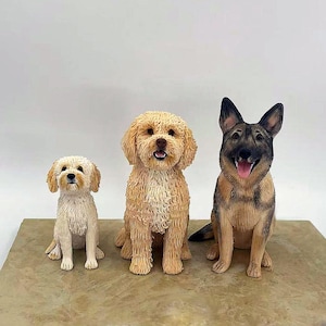 May include: Three dog ornaments: a small cream-coloured dog, a medium-sized golden doodle, and a German Shepherd. The dogs are sitting on a brown surface against a white background. The German Shepherd has its tongue out.