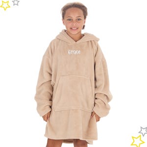 May include: A tan oversized hoodie with a front pocket and hood. The word "Grace" is embroidered in white on the chest. The garment appears to be made of a soft, plush material. The person is wearing the hoodie.