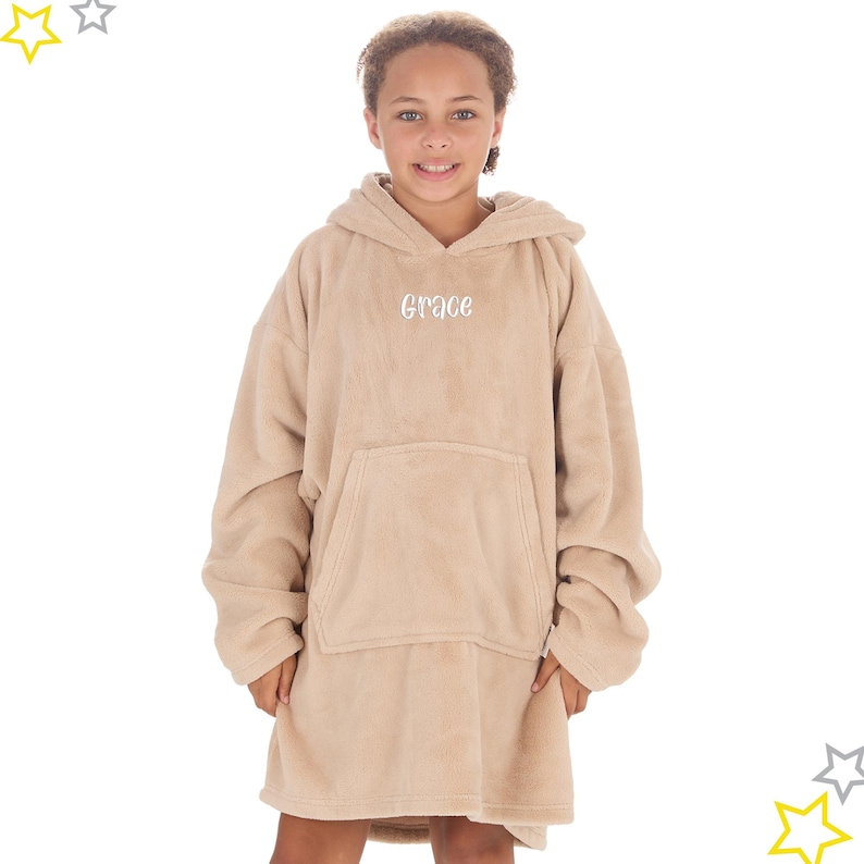 May include: A tan oversized hoodie with a front pocket and hood. The word "Grace" is embroidered in white on the chest. The garment appears to be made of a soft, plush material. The person is wearing the hoodie.