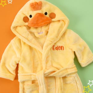 Puede incluir: Batista amarilla con capucha con un dise&ntilde;o de pato en la capucha. La bata tiene un cintur&oacute;n de atar y un bolsillo. La bata est&aacute; personalizada con el nombre "Eden".