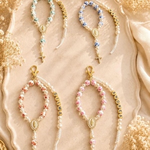 May include: Four personalized rosary keychains with pearl beads, gold-tone accents, and cross pendants. Each rosary features a different color scheme: white and green, white and blue, white and red, and white and pink. Names are spelled out in gold beads.
