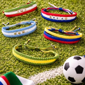 May include: A collection of colorful woven bracelets, each featuring the colors of different national flags, including USA. The bracelets are displayed on a green surface with a soccer ball and a flag-themed ribbon.