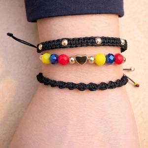 May include: Three bracelets on a wrist. The top bracelet is black with gold beads. The middle bracelet has red, yellow, and blue beads with a gold heart charm. The bottom bracelet is black with a knotted design.