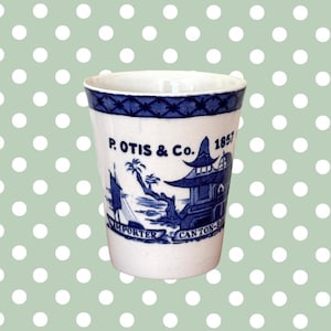 May include: A white ceramic cup with a blue and white design. The cup has a blue band around the top and a blue and white scene of a pagoda and a boat on the side. The text "P. OTIS & Co. 1857 IMPORTER CANTON-CHINA" is printed on the cup.