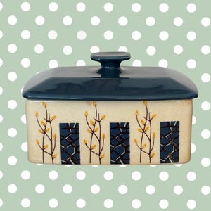 May include: A rectangular ceramic butter dish with a navy blue lid and handle. The off-white base features a repeating pattern of blue rectangles with a textured design and yellow leaf-like accents. The dish is designed for kitchen use.